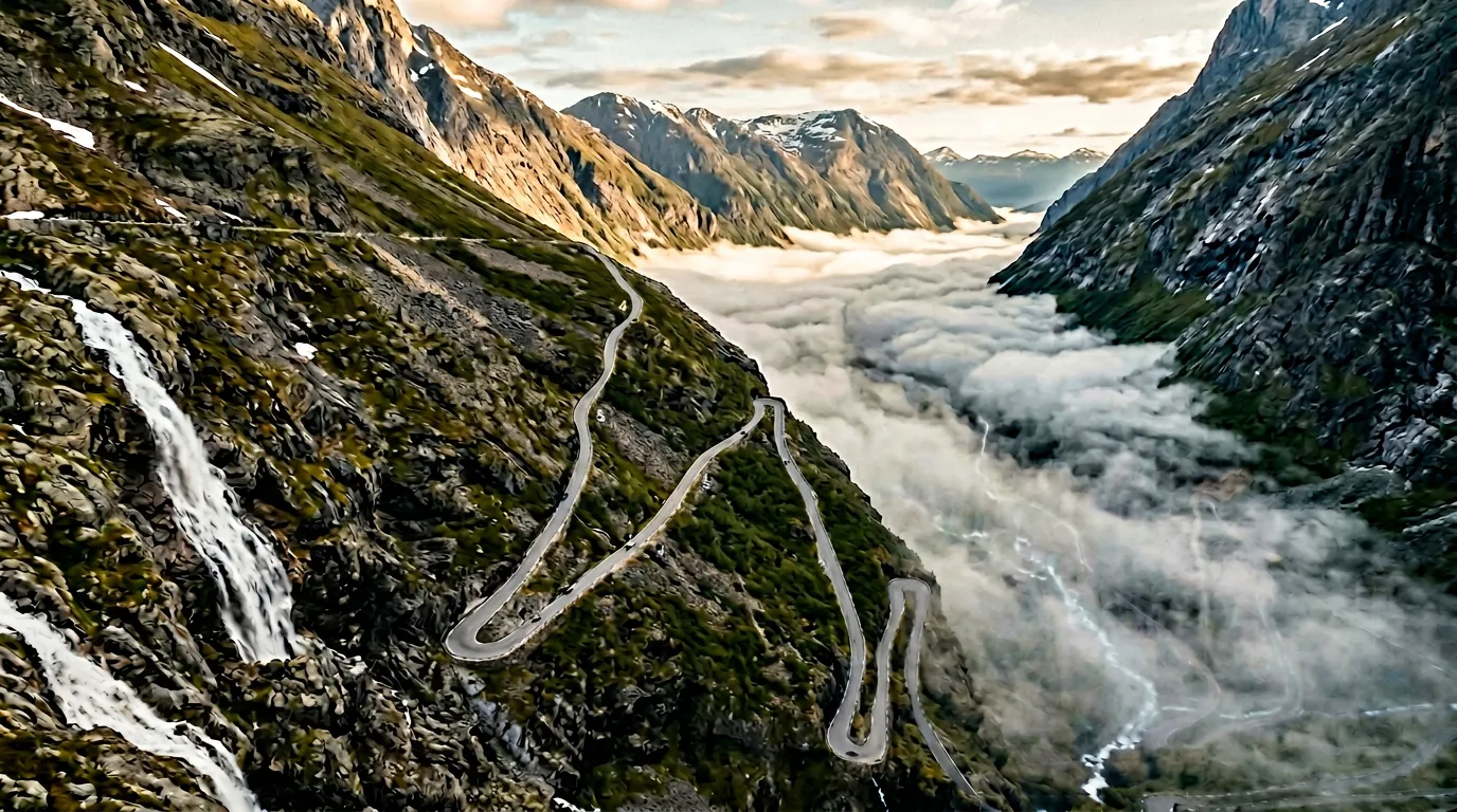 Aerial view of Trollstigen's eleven hairpin turns climbing the steep mountain wall, morning mist filling the valley below, Stigfossen waterfall visible as a white ribbon beside the road